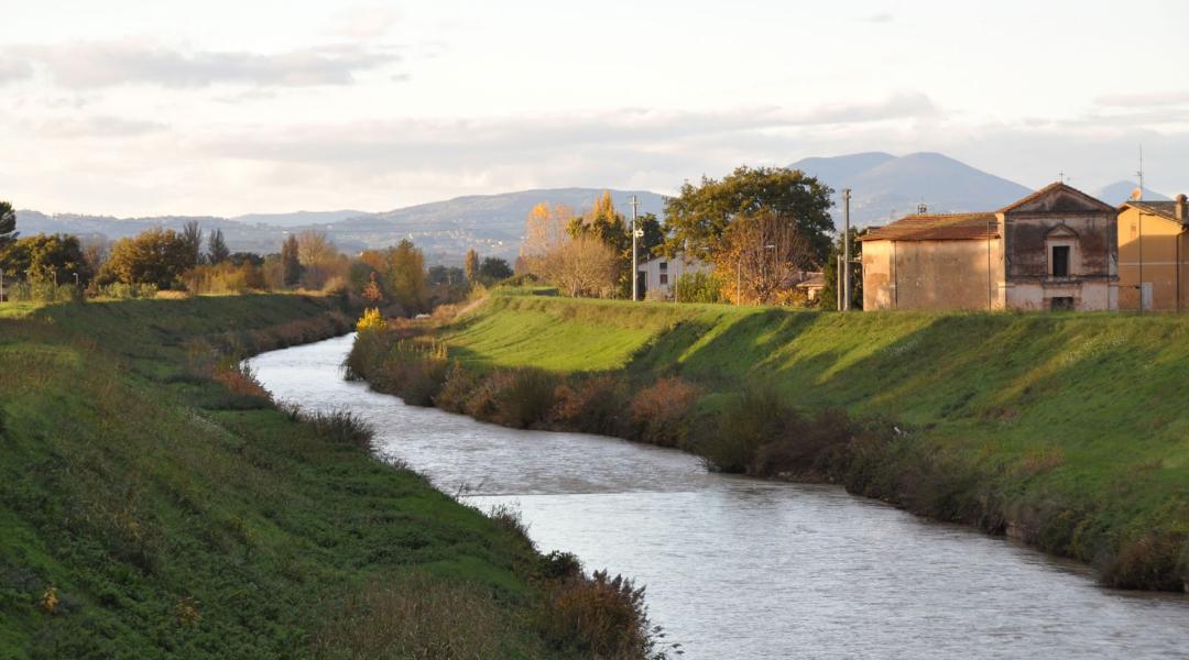 River flowing between the green banks of Cannara, with rural houses and hills lit by the sunset.