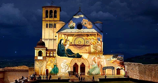  Facade of the Basilica of Saint Francis in Assisi illuminated at night with artistic projections depicting Nativity scenes, observed by visitors on the lawn in front. 